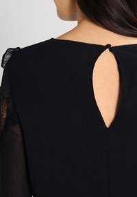 Námořnická blůza s texturovanou tkaninou má kulatý výstřih a klíčovou vykrojení na zadní straně, zdůrazněnou volánovými rukávy.