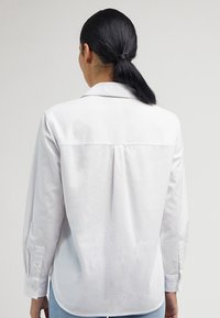 Chemise blanche à manches longues en coton avec un col pointu, un empiècement au dos et des poignets boutonnés. Elle présente une coupe décontractée et une texture lisse.