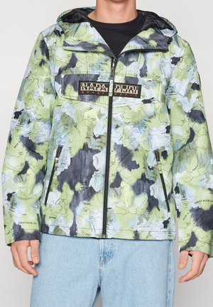 Man wearing green, black, and white map-patterned zip-up jacket with hood and two zipped side pockets, paired with light blue jeans.