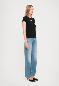 Vrouw die staat en een zwart T-shirt met korte mouwen, lichtblauwe wijde jeans en zwarte puntige hakken draagt, op een witte achtergrond.