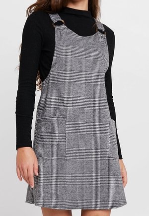 Femme portant une robe tablier à carreaux gris avec des bretelles à anneaux argentés sur un pull col roulé noir à manches longues.