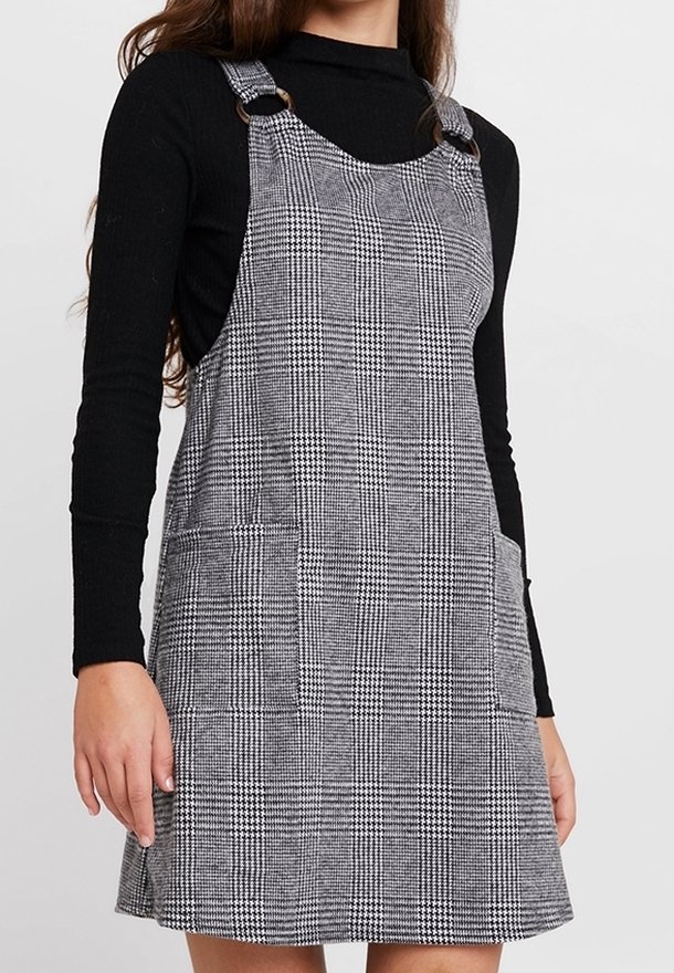 Femme portant une robe tablier à carreaux gris avec des bretelles à anneaux argentés sur un pull col roulé noir à manches longues.