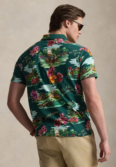 Άνδρας με πράσινο πόλο πουκάμισο με πολύχρωμα λουλουδένια και μοτίβα παίκτη πόλο, μπεζ παντελόνι και σκούρα γυαλιά ηλίου, στέκεται μπροστά σε ουδέτερο φόντο.