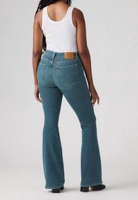 Jeans acampanados de talle alto en color verde azulado con un diseño distintivo en el bolsillo trasero y una etiqueta de cuero marrón, combinados con una camiseta sin mangas blanca.