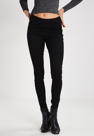 Femme portant un pantalon noir ajusté, des bottines noires et un pull court gris foncé côtelé, debout les mains sur les hanches.
