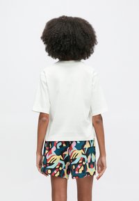 Biele tričko s krátkym rukávom a rovnostrižným strihom v kombinácii s viacfarebnými šortkami s abstraktnými vzormi v žiarivých farbách.