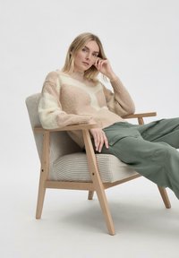 Lichtbruin en crème geblokt patroon trui met een fluffy textuur, gecombineerd met losse groene cargobroek, zittend in een licht houten armstoel met ribbelstof.