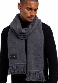 Écharpe en tricot grise avec des extrémités frangées et un motif texturé, arborant une étiquette noire avec le logo "FALKE", portée sur un pull noir.