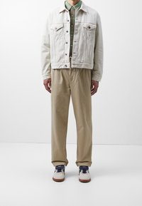 Mand iført en off-white denimjakke, grøn skjorte med krave, beige løstsiddende bukser og hvide sneakers med blå og grønne detaljer.