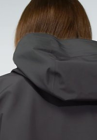 Person with straight brown hair wearing a black hooded jacket viewed from the back against a plain background.