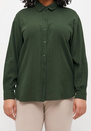 Kvinde iført en langærmet, knappet, mørkegrøn tekstureret skjorte med beige bukser, stående foran en ensfarvet baggrund.