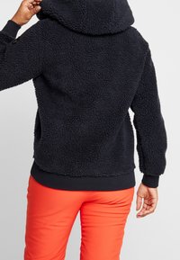 Marinefarbener Fleece-Pullover mit strukturierter Oberfläche, Kapuze und gerippten Bündchen. Kombiniert mit leuchtend orangefarbener Hose für einen Kontrast.