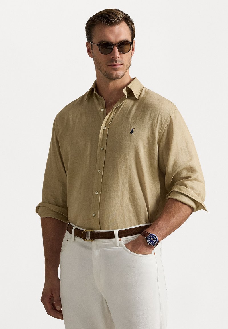 Camisa de linho bege com botões e mangas enroladas, detalhe do pequeno logotipo. Combinada com calças brancas e cinto castanho, complementada por óculos de sol escuros.