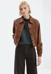 Giacca corta in pelle marrone con collo a risvolto, chiusura con bottoni e polsini a coste, indossata sopra un maglione scuro e pantaloni a vita alta.