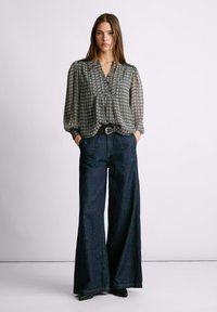 Blusa a maniche lunghe, a motivi bianco e nero con scollatura a V e dettagli plissettati, abbinata a jeans scuri a gamba larga e una cintura nera.