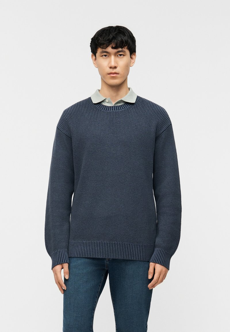 Νεαρός άνδρας που φοράει σκούρο μπλε πλεκτό πουλόβερ πάνω από ανοιχτό γκρι πουκάμισο με γιακά και μπλε τζιν, στέκεται μπροστά σε απλό λευκό φόντο.