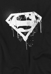 Black t-shirt featuring a large, distressed white Superman logo with paint splatters and drips. Soft texture, casual design.