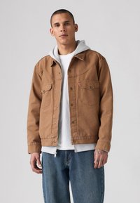 Bruine denimjack met twee borstzakken, zilverkleurige hardware en een grijze capuchontrui eronder, gecombineerd met blauwe jeans.