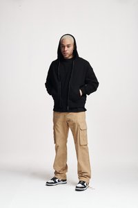 Zwarte zip-up hoodie met zakken, gecombineerd met kaki cargobroek met zijzakken en zwart-witte sneakers. Minimalistische achtergrond.