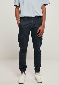 Homme portant un pantalon jogger cargo noir et des baskets blanches, debout les mains dans les poches devant un fond uni.