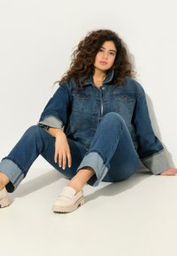 Tummansinisestä farkkutakista ja -farkuista, joissa on vaaleansinisiä yksityiskohtia. Pyöreät beige loaferit paksulla pohjalla. Malli istuu valkoista taustaa vasten.