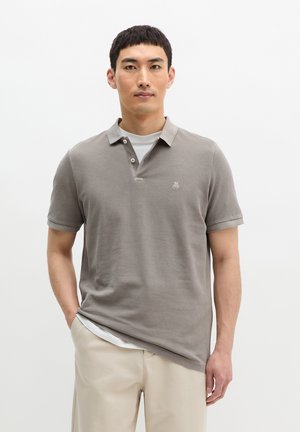 Uomo con capelli neri corti che indossa una polo grigia sopra una maglietta bianca e pantaloni beige, in piedi davanti a uno sfondo bianco uniforme.