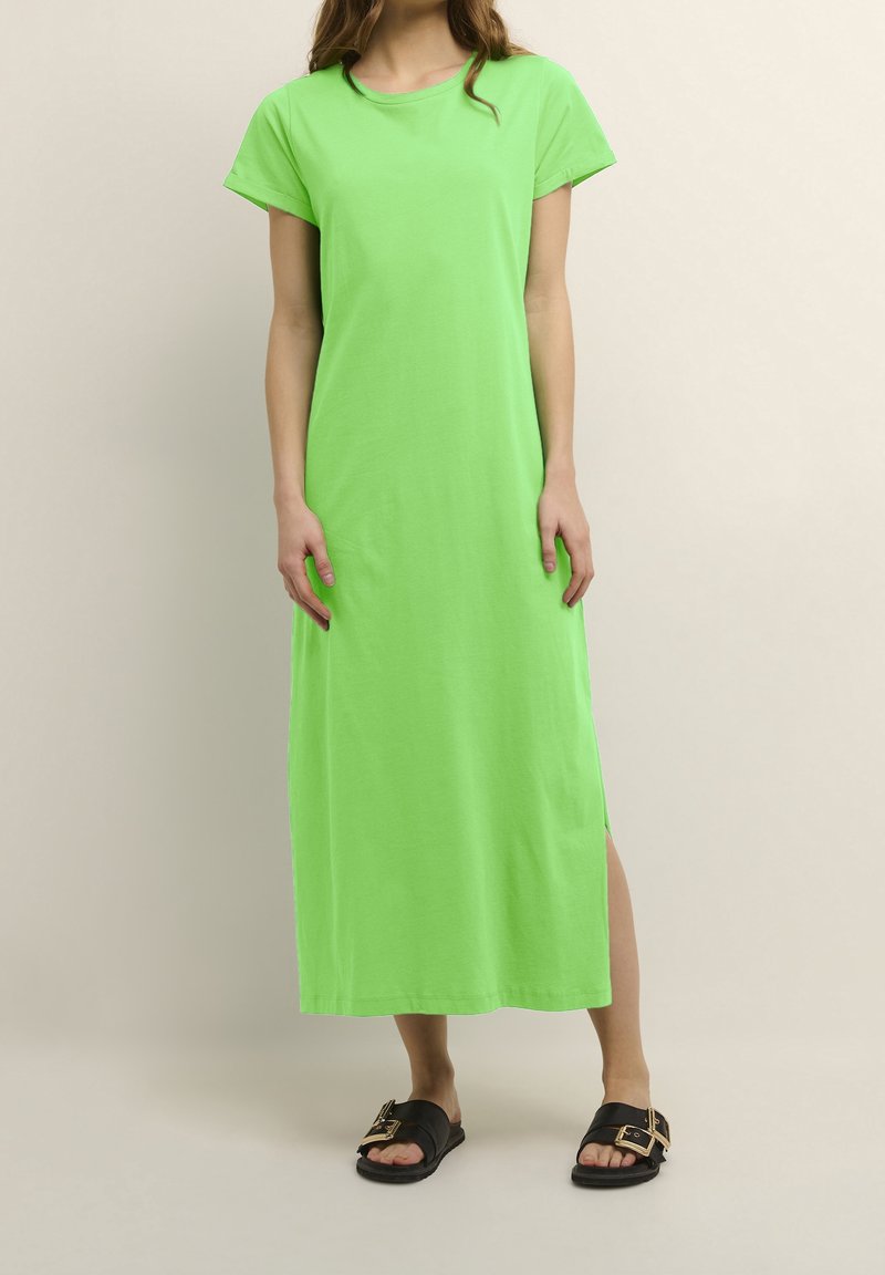 Femme portant une robe verte vif à manches courtes, longue jusqu'aux chevilles avec une fente latérale, et des sandales noires à enfiler avec des boucles dorées.