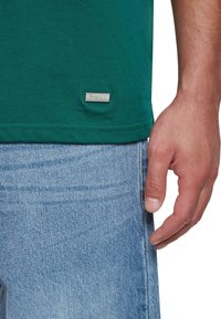 Grøn bomulds T-shirt med en glat tekstur, der har et lille sølvlogo-tag i kanten; parret med lyseblå denimjeans.