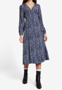 Langärmliges Wickelkleid in Marineblau mit Schlangenprint, V-Ausschnitt, mittellanger Länge und strukturiertem Stoff. Kombiniert mit schwarzen Stiefeln.