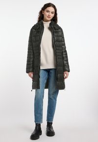 Donkergroene pufferjas met gewatteerd patroon, lange mouwen en een hoge kraag. Gecombineerd met een beige trui en blauwe jeans, gedragen met zwarte enkellaarsjes.