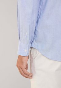 Camisa de manga larga azul claro con puños abotonados, que presenta una textura sutil y un corte ajustado, combinada con pantalones de color claro.