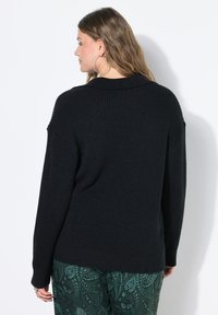 Vrouw met lang haar in een zwarte geribbelde trui en groene paisley-gepatroonde broek, met de rug naar de kijker tegen een witte achtergrond.