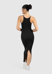 Vestido maxi negro sin mangas con una silueta ajustada, que presenta aberturas laterales y una textura suave. Llevado con zapatillas deportivas blancas.