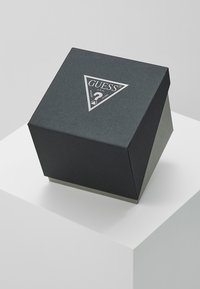 Caja cuadrada texturizada negra con una tapa en forma de pirámide, que presenta un logo plateado en un triángulo en la parte superior y una base gris sutil.