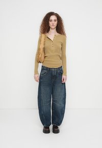 Mladić s kovrčavom kosom nosi žutu majicu dugih rukava, široke tamne traperice, tamne cipele i nosi teksturiranu smeđu torbu preko ramena.