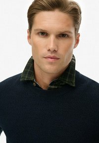 Marineblauer strukturierter Pullover mit rundem Halsauschnitt, über einem grün-schwarz karierten Hemd mit Kragen getragen, das Knöpfe und sichtbare Kragendetails aufweist.