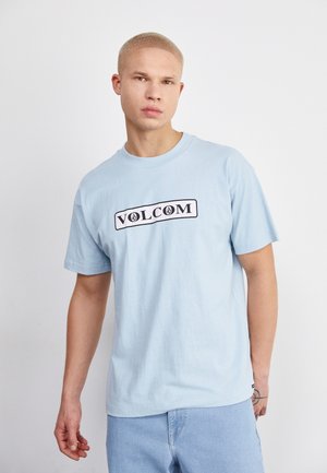 Camiseta de algodón azul claro con un logotipo negro de "VOLCOM" dentro de un borde rectangular en el frente. Cuello redondo y mangas cortas.