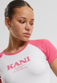 T-shirt à manches courtes avec des manches raglan roses, corps blanc, arborant le logo "KANI" en texte rose sur le devant. Matière en coton lisse.
