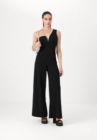 Svart jumpsuit med dyp V-hals og én skulder. Vid bendesign, glatt tekstur, accessorert med en liten blank veske og klumpete platåsko.