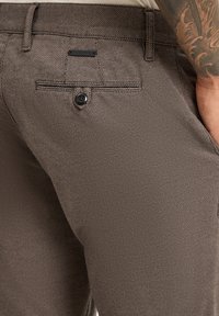 Braune, strukturierte Hose mit dezentem Muster, ausgestattet mit einer Gesäßtasche mit Knopf und einem kleinen schwarzen Logo-Akzent.