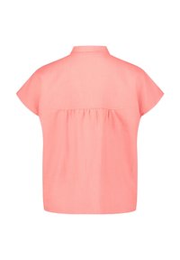 Takakuvassa lyhythihainen korallinpunainen pusero, jossa pieni kaulus ja poimutettu kangasosa yläosan alapuolella.