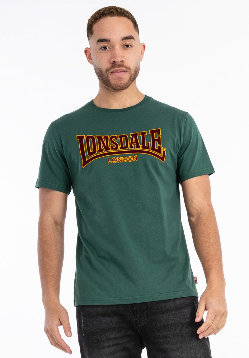 Grøn bomulds T-shirt med "LONSDALE" og "LONDON" trykt med fede gule og røde bogstaver. Klassisk rund hals og korte ærmer.