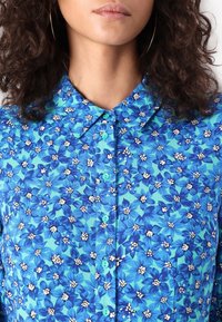 Blumenhemd mit Knopfleiste in hellblau mit weißen und dunkelblauen Blumenmustern. Mit klassischem Kragen und türkisfarbenen Knöpfen.