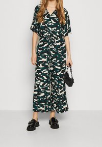 Femme portant une combinaison à motif abstrait vert, noir et beige, tenant un petit sac à main noir et portant des mocassins compensés noirs.