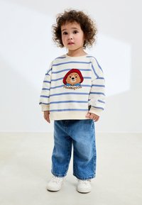 Sweat-shirt rayé blanc et bleu avec un ours Paddington brodé et le texte « Paddington », associé à un jean bleu ample et des baskets blanches.