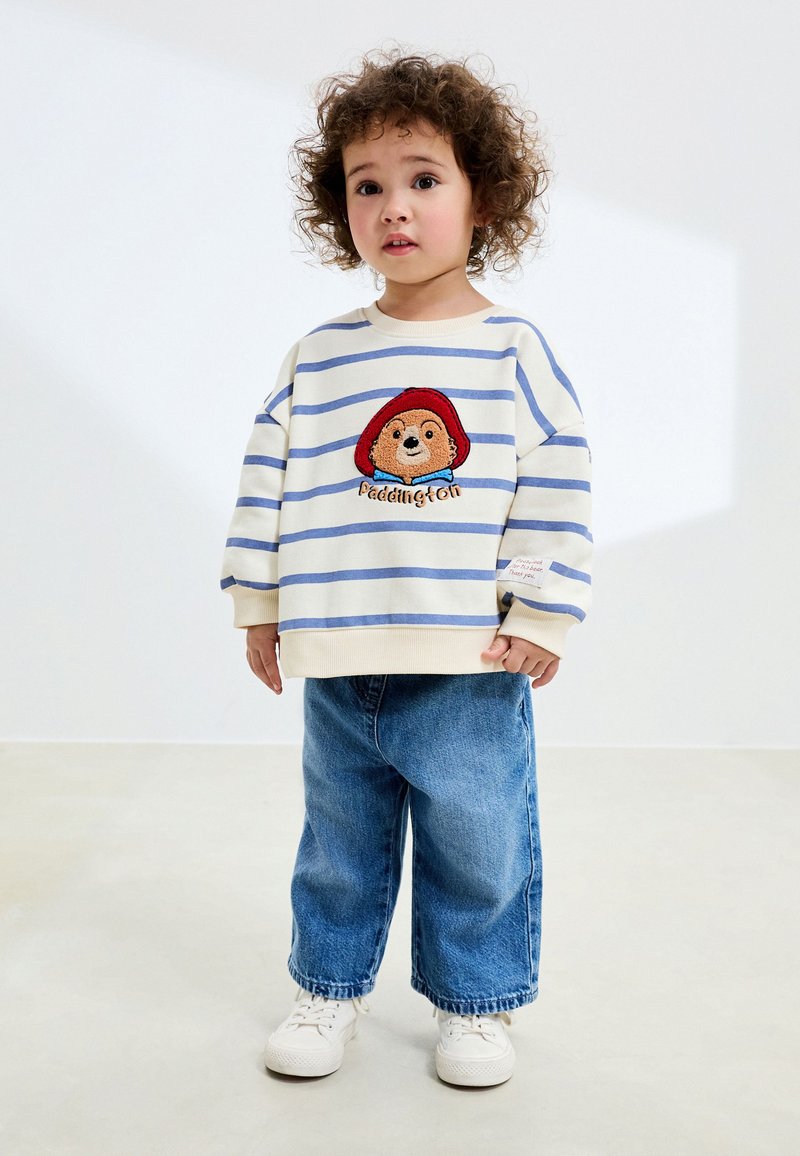 Sweat-shirt rayé blanc et bleu avec un ours Paddington brodé et le texte « Paddington », associé à un jean bleu ample et des baskets blanches.