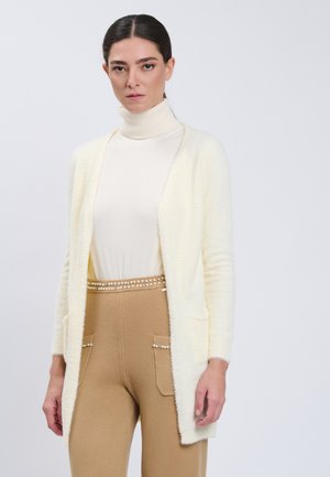Mujer con cuello alto blanco y cárdigan crema de textura suave con pantalones camel que presentan adornos de perlas en los bolsillos y la cinturilla.