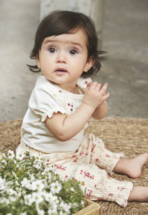 Chemise crème à manches courtes avec des bords froncés, assortie à un pantalon léger à motif floral. Bébé assis sur un tapis tissé près de fleurs blanches.
