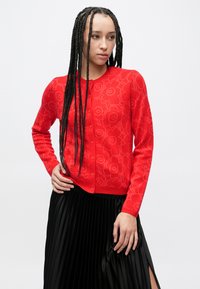 Rode cardigan met bloemenpatroon, knoopsluiting, lange mouwen en geribde zoom. Gecombineerd met een zwarte geplooide rok met een zijsplit.