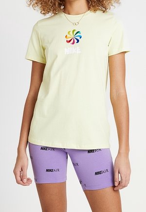 Kobieta ubrana w jasnożółtą koszulkę Nike z wielokolorowym spiralnym logo oraz fioletowe rowerowe spodenki Nike Air z czarnymi wzorami tekstowymi.